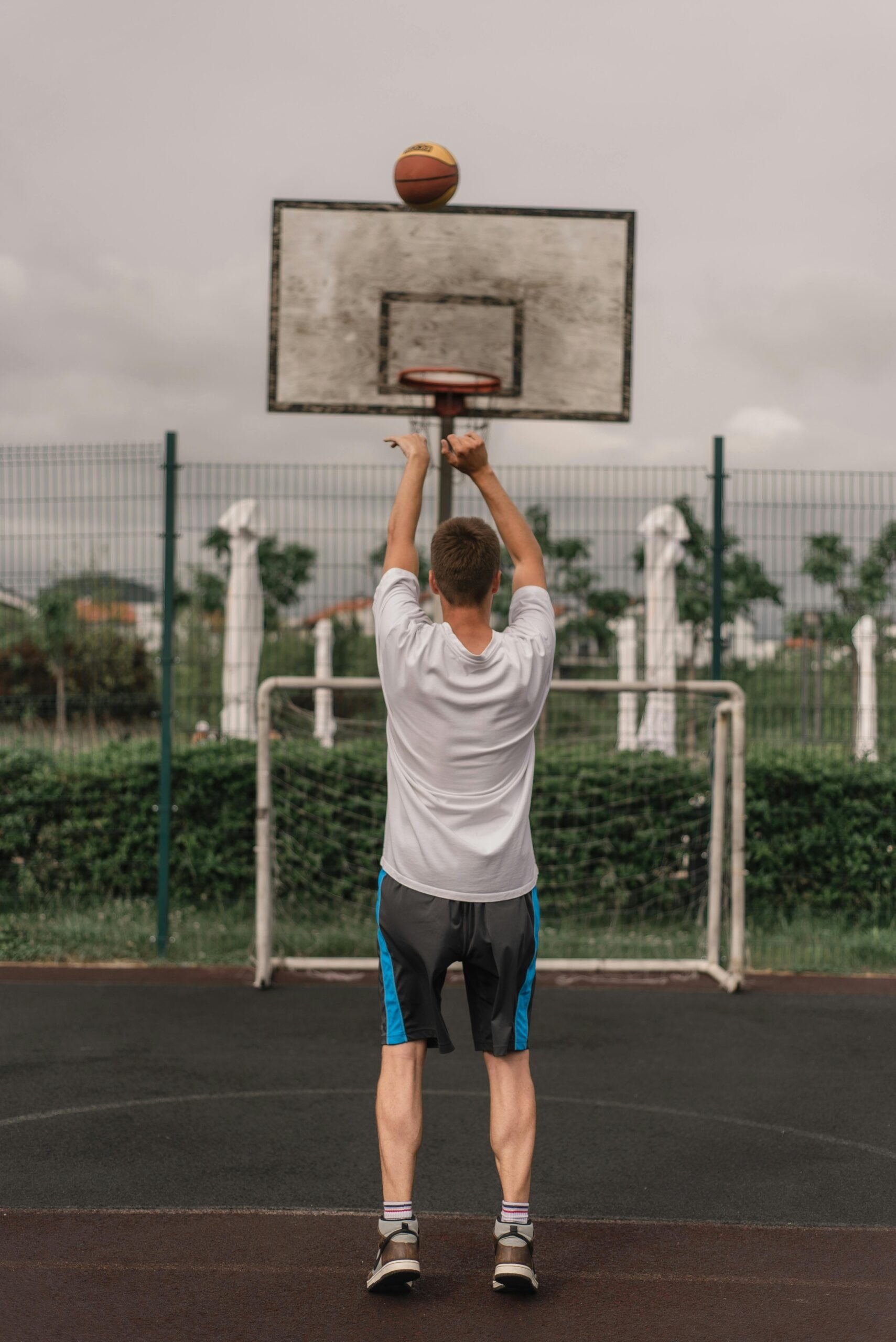 Back view of a young man playing basketball, shooting towards the hoop on an outdoor court.